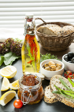 Dried tomatoes in jar and bottle of olive oil with spices, mussels and black olives and in ceramic bowls, bread, fresh tomatoes, sliced lemon, herbs on black stone board. Mediterranean food