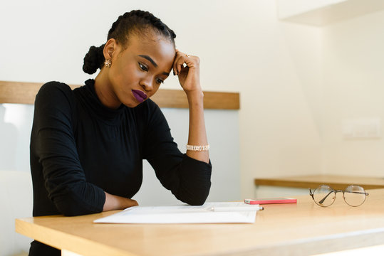 Thoughtful worried African or black American woman holding her forehead with hand looking at notepad in office