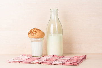 Faceted glass of milk with a bun on it stands on a red squared kitchen towel near a traditional opened old fashioned bottle of milk on light wooden textured background.