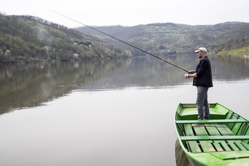 Naklejka premium Side View Of Sport Fisher With Cap And Vest Fishing From Green Canoe And Enjoying His Vacation On Beautiful Lake 