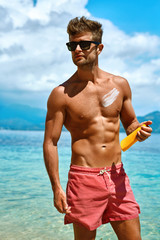 Sun Skin Protection In Summer. Handsome Man In Sunglasses Sunbathing With Sunscreen Lotion On Sexy Athletic Muscular Body. Male Fitness Model Tanning Using Solar Block Cream For Healthy Tan. Skincare 