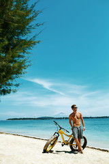 Fitness Male Model With Bike Sunbathing On Sun, Enjoying Summer Travel Vacation. Handsome Sexy Man In Sunglasses With Muscular Body, Healthy Tan Skin Tanning Near Bicycle On Sand Beach. Summertime 