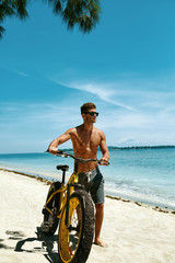 Fototapeta na wymiar Summer Beach Sport. Athletic Man With Muscular Body Riding Yellow Sand Bicycle At Tropical Seaside. Fitness Male Model With Bike On Holiday Travel Vacation. Sporting Activity, Active Lifestyle Concept