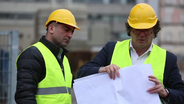 Two male engineers wearing safety jackets and hardhats checking technical drawings
