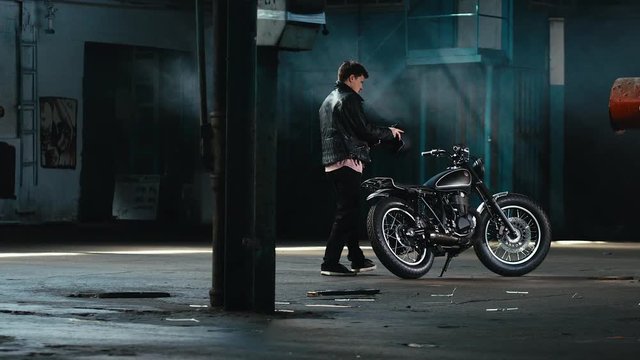 Young thoughtful Caucasian male biker in leather jacket mounts his custom cafe racer motorcycle in large warehouse garage. 60 FPS slow motion Blackmagic URSA Mini RAW graded footage