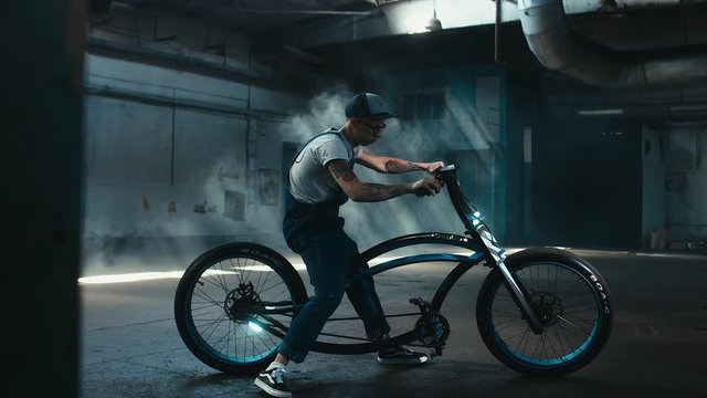 Young Caucasian male in denim overalls with tattoos riding his custom made bicycle in large warehouse garage. 60 FPS slow motion Blackmagic URSA Mini RAW graded footage