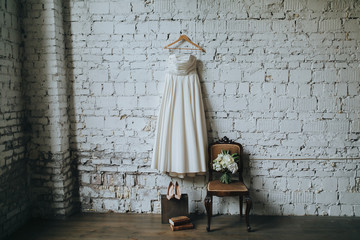 white wedding dress, shoes and a bouquet of white flowers and green against a white brick wall in loft