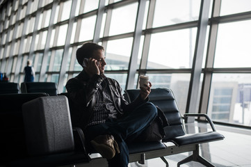 Portrait of young handsome man wearing casual style clothes sitting on the bench in modern airport using smartphone. Passenger travelling with luggage bag making call, while waiting for his flight