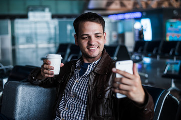 Portrait of a young man laughing and taking selfie with coffee at the airport waiting his flight
