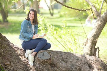 Happy young female student is reading a book in a park. She is sitting on a tree, looking away and smiling.