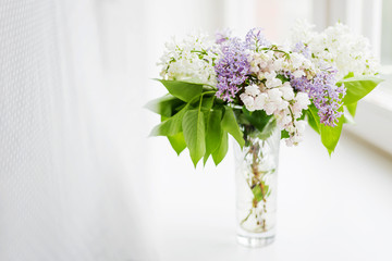 Lilac flowers in glass vase on window sill. Natural spring background with white and violet flowers.