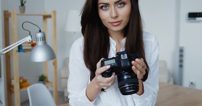 Attractive smiling brunette female photographer in white shirt checking an image holding her camera, office interior at background.
