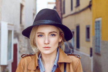 Close up of beautiful young blonde woman in old town street. Wearing black fashion hat, coat and shirt. Her hair is tied to ponytail. 