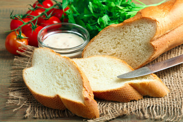 sliced baguette with greens and a knife with sauce on sackcloth on wooden background with tomatoes