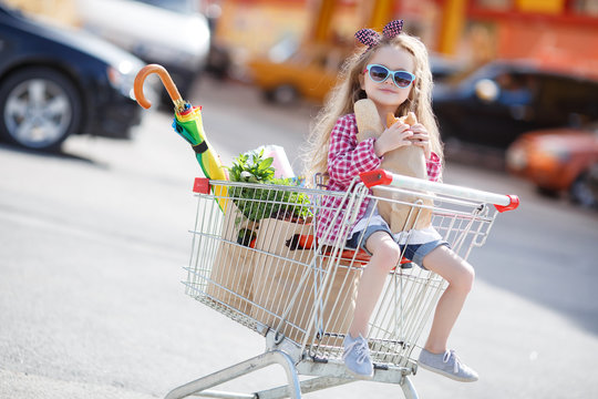 Little girl,with thick blonde long hair,dressed in a plaid shirt and denim shorts,on his head a black-and-red bow,wears sun glasses,sitting in the shopping cart on wheels near a large supermarket