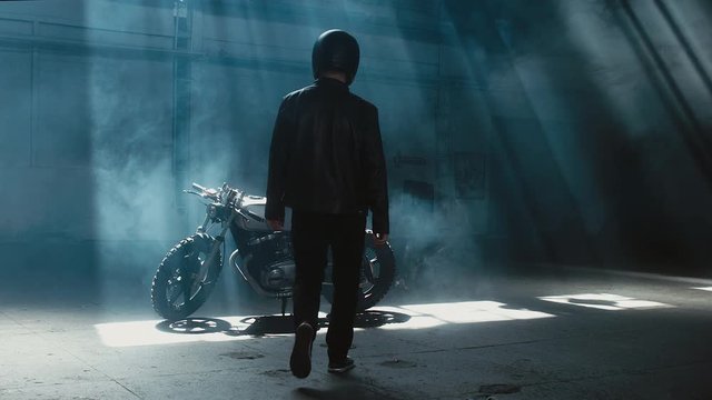 Young thoughtful Caucasian male biker in leather jacket mounts his custom cafe racer motorcycle in large warehouse garage. 60 FPS slow motion Blackmagic URSA Mini RAW graded footage
