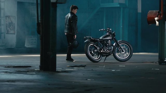 4K cinemagraph - seamless loop - young thoughtful Caucasian male biker in leather jacket standing near custom cafe racer motorcycle in large warehouse garage with helmet in hands looking forward