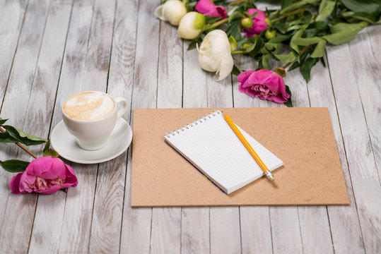 Notebook with a pencil next to coffee and peonies flowers on wooden background.