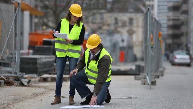 Crouched engineer checking blueprints with a female colleague on construction site