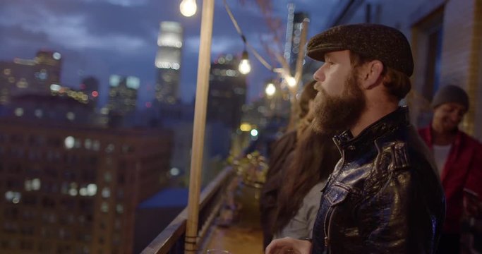 Bearded man in leather jacket and flat cap looks around from balcony then drinks beer in Downtown Los Angeles, California. Other guests in background.  Recorded in slow motion at 60fps.