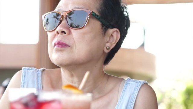 Beautiful Asian senior woman wearing sunglasses relax with cocktail on beach vacation 