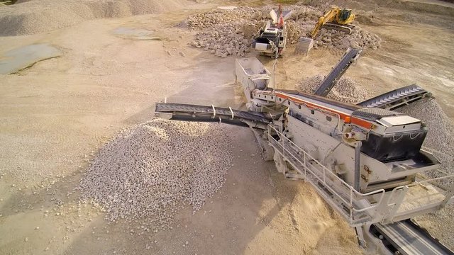 The big trucks and machineries in the quarry area. There are limestone crusher and the backhoe surrounded with big stones