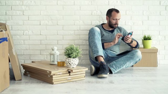 Young man using tablet and smartphone while sitting on floor at his new home
