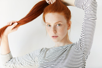 Youth and skin care concept. Portrait of pretty young female with red hair and clean fresh skin with freckles, wearing stylish striped top, touching hair, looking with serious expression at the camera