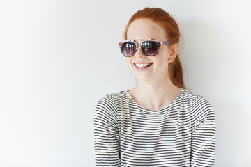 Headshot of young Caucasian redhead female office worker or student relaxing and having fun with her friends indoor. Hipster teenager wearing stylish striped top looking away with happy expression