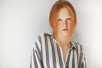 Headshot of cute Caucasian redhead freckled teenager model in striped night suit, looking at the camera with serious and thoughtful expression, ready for going to bed. Youth and skin care concept.