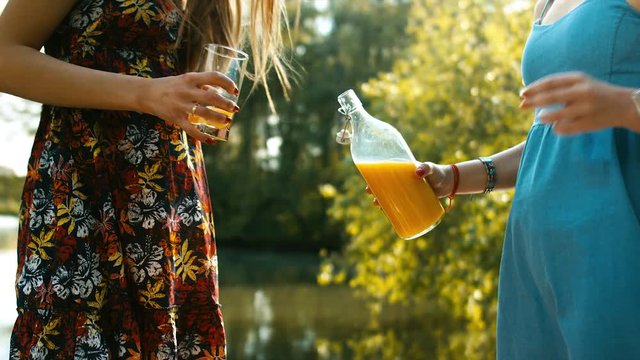 Two happy young Caucasian teen drinking orange juice near a lake. Summer holidays, friendship concept. 60 FPS slow motion shot. Blackmagic URSA Mini RAW graded footage