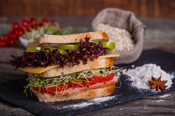 Sandwich with cereals bread with lettuce, slices of fresh tomatoes and sweet pepper, ham, prosciutto, salami, cheese  and  onion sprouts on dark marble background