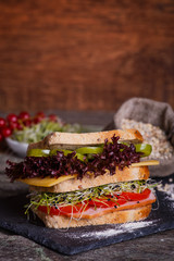 Sandwich with cereals bread with lettuce, slices of fresh tomatoes and sweet pepper, ham, prosciutto, salami, cheese  and  onion sprouts on dark marble background