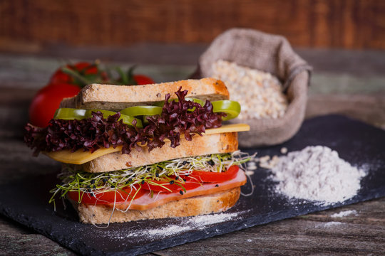 Sandwich with cereals bread with lettuce, slices of fresh tomatoes and sweet pepper, ham, prosciutto, salami, cheese  and  onion sprouts on dark marble background