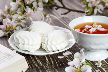 Cup of tea with vanilla zephyr and a book on the old wooden table. Petals and flowers of cherry and apple trees in the spring garden. Rest and relaxation