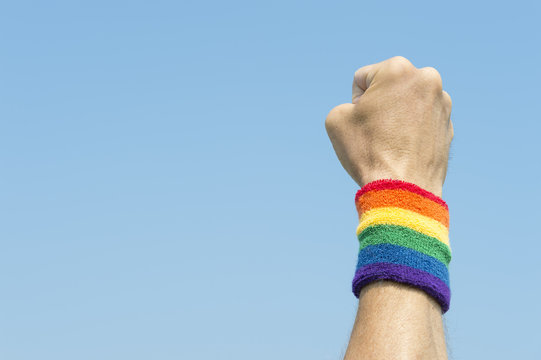 Gay athlete punching the air with gay pride rainbow colors wristband against bright blue sky