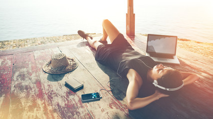 Young woman with laptop listening music with headphones near the ocean. Lens flares effect,...