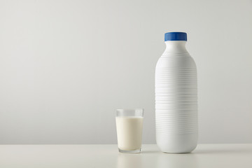 Transparent glass with fresh organic milk near plastic riffled blank bottle with blue cap isolated on side of white table Space for your text above Retail set, rich texture