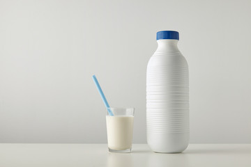 Transparent glass with fresh organic milk and blue drinking straw inside near plastic riffled blank bottle with blue cap isolated on side of white table Space for your text above Retail set