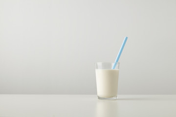 Transparent glass with fresh organic milk and blue drinking straw inside isolated on side of white table Space for your text above