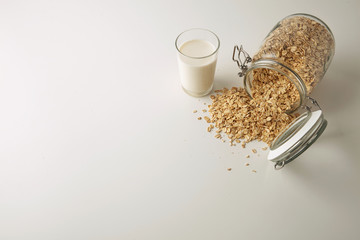 Transparent glass with fresh organic milk near lying opened rustic jar with rolled oats spread out isolated in center on white table top side view