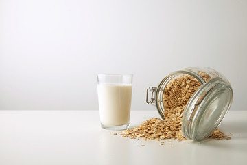Transparent glass with fresh organic milk near lying half opened rustic jar with rolled oats spread...