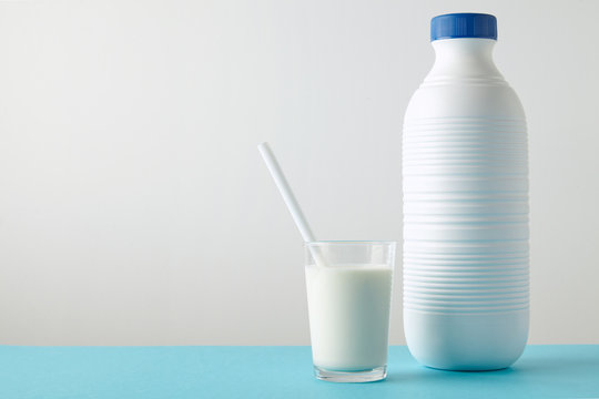 Transparent glass with fresh organic milk and white drinking straw inside near blank riffled plastic bottle with blue cap isolated on pastel blue table Space for your text above rich texture