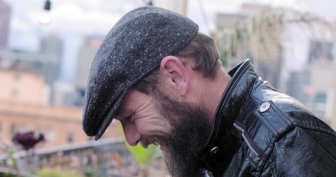 Bearded man in leather jacket and flat cap holds beer glass, talks and laughs at a party in Downtown Los Angeles, California.  Recorded in slow motion at 60fps.
