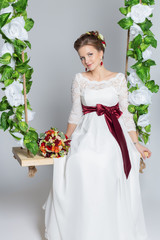 beautiful lovely bride is sitting on a swing with a beautiful bouquet of colorful flowers in a white dress with evening hairstyle