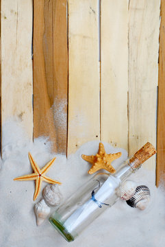Summertime image with beachy theme of clean white sand scattered on boardwalk planks with starfish, seashells and a message in a bottle making a lower border for vertical image