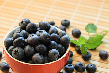 Blueberry in Red Bowl. Mint Leaves and Scattered Berries on Straw Surface.