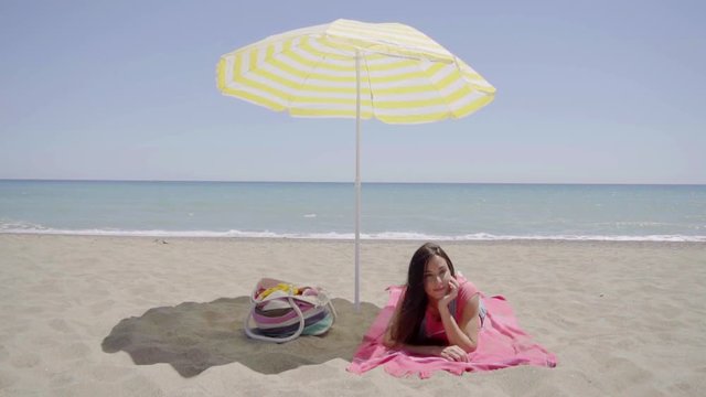 Single Cute Young Lady Laying Down On Beach Blanket Next To Bag In Shade Under Yellow Umbrella With Ocean In Background