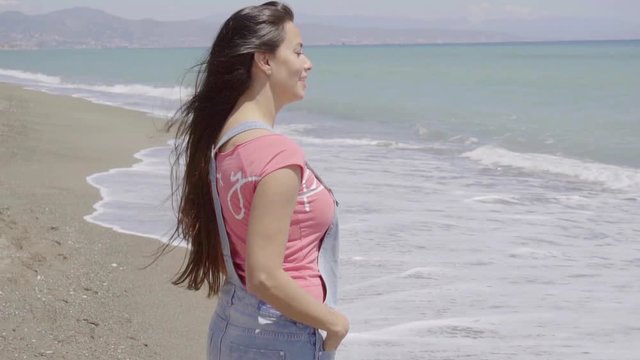 Front view on cheerful young woman in long brown hair  pink top and blue jeans shorts walking along beach with mountains behind her