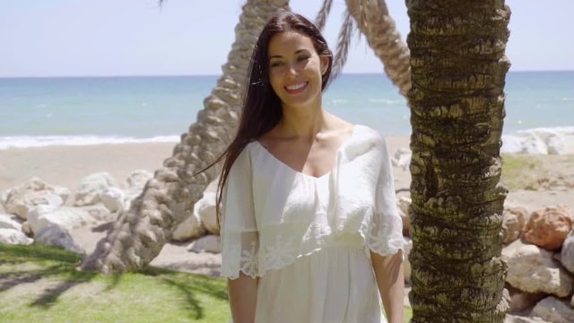 Cheerful single gorgeous woman wearing white flowing blouse standing near palm trees with ocean in background